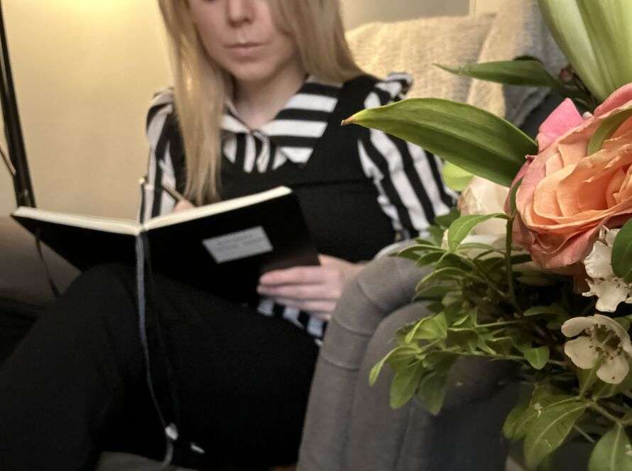 Natalie Forslind sitter och skriver i sin svarta dagbok. Blommor syns i förgrunden på bilden.